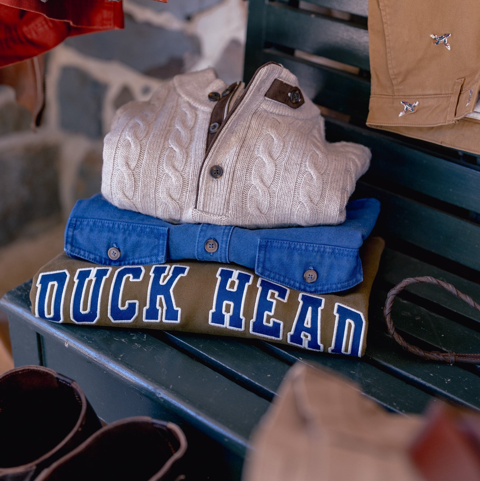 A stack of folded Duck Head brand shirts on a dark green wooden bench, featuring a cream cable-knit shirt on top, followed by a blue button-down, a tan button-down with blue lettering, and a dark shirt underneath. A red jacket hangs on the left, and a tan shirt is draped over the bench back.
