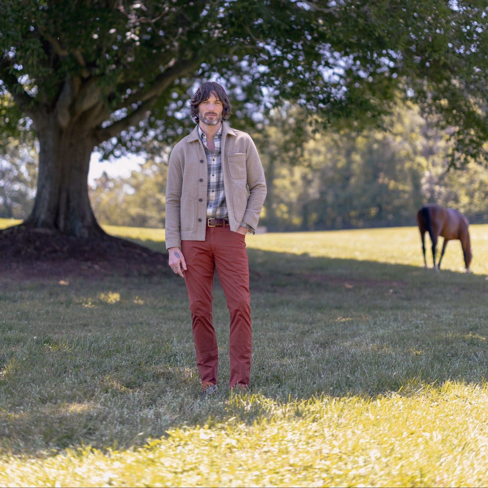 A man wearing a beige jacket, brown shirt, and rust-colored pants stands in a grassy field under a large tree, with a horse visible in the background.