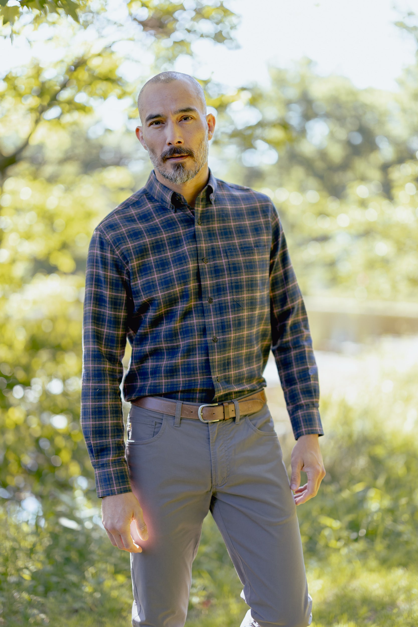 A man wearing a long-sleeve plaid button-down shirt in navy, blue, and yellow, paired with gray pants and a brown belt, standing outdoors in a sunlit, green wooded setting.
